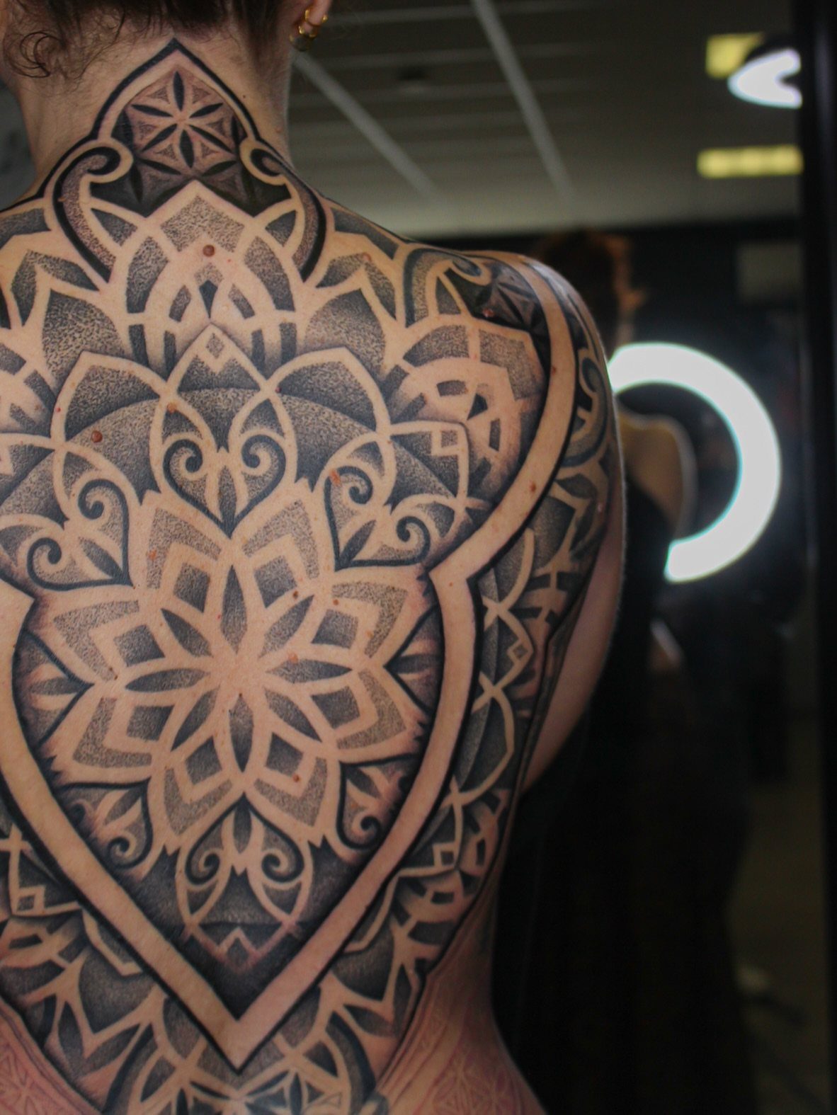 Intricate black and grey mandala tattoo covering the upper back and spine of a woman.