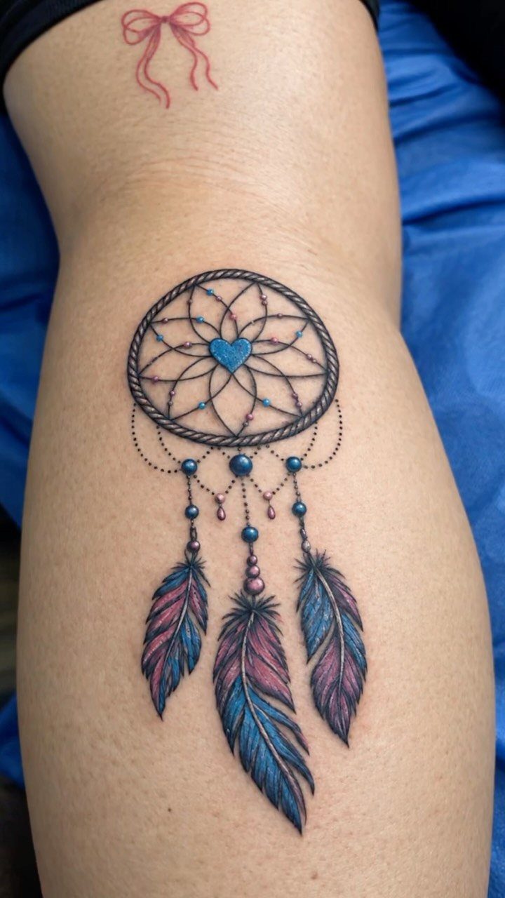 Dreamcatcher tattoo with a blue heart and colorful feathers on a person's leg
