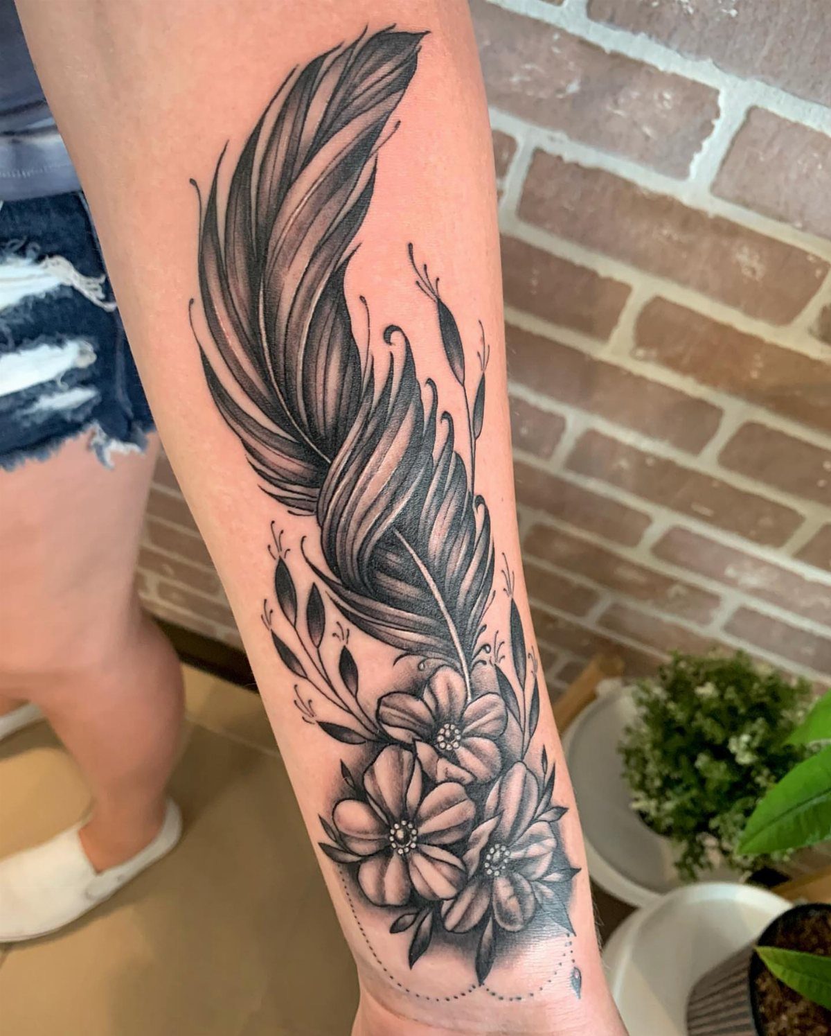 Detailed black and grey tattoo of a feather and flowers on a forearm.