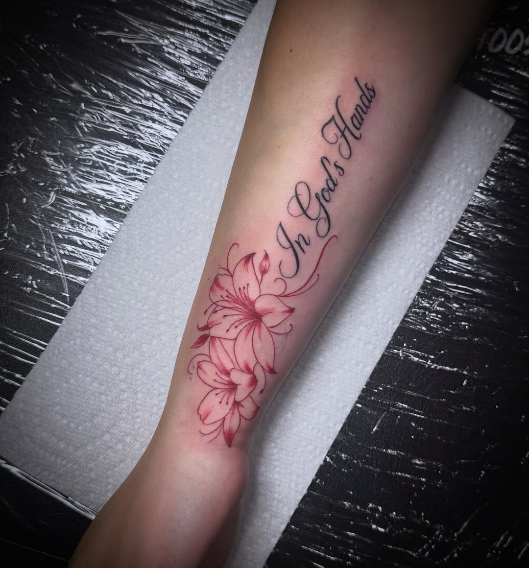 Forearm tattoo of pink lilies and the cursive phrase In God's Hands.