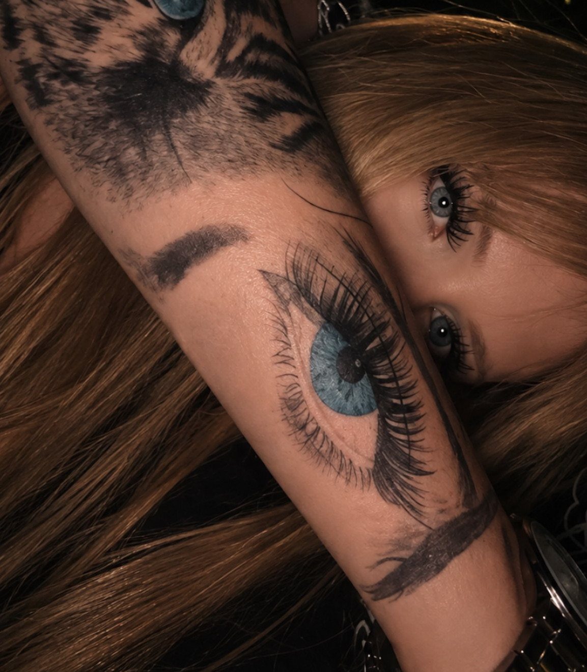 Realistic blue eye tattoo on a forearm next to a person's actual eye