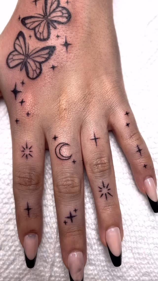 Hand with small black ink tattoos of butterflies, a crescent moon, and stars.
