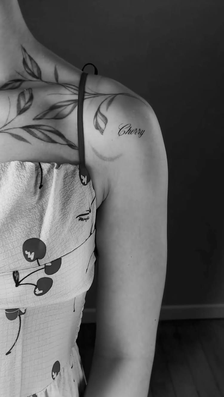 Woman's shoulder with a cursive Cherry tattoo and leaf branch collarbone tattoo.