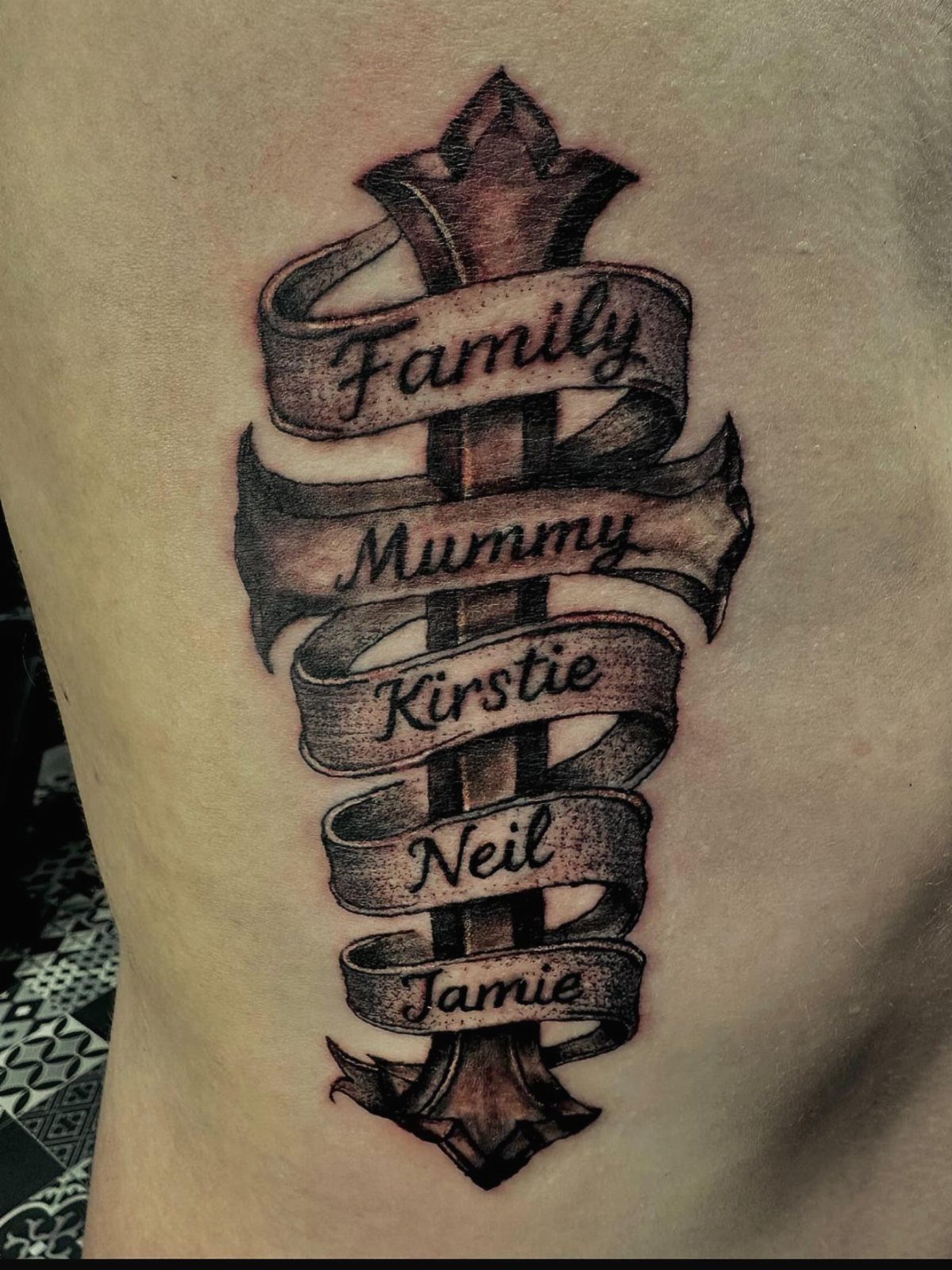 Rib tattoo of a sword wrapped in a ribbon with family names.