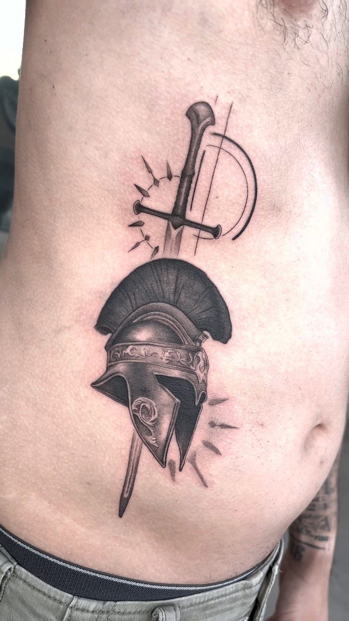 Spartan helmet and sword tattoo on a man's ribcage in black and grey ink