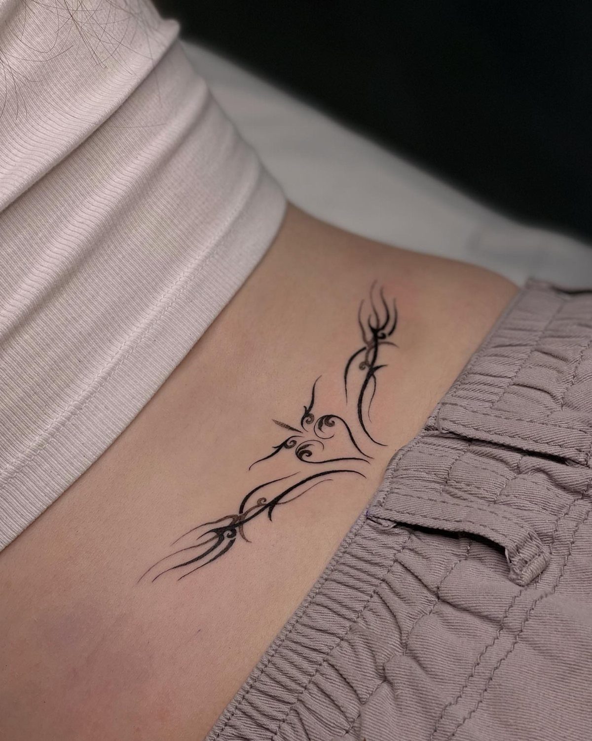 Tribal tattoo on a person's ribcage between a white shirt and grey pants.