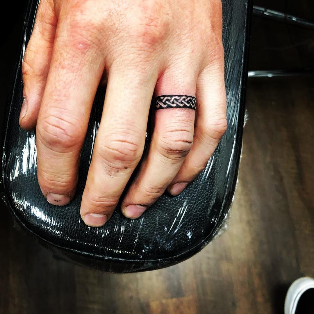 Braided Celtic knot tattoo encircling a person's ring finger on a black armrest