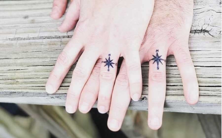 Two ring fingers with matching black compass rose tattoos resting on wooden planks.