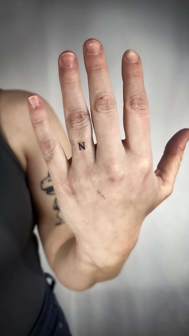 Small black letter N tattoo on the ring finger of a hand.