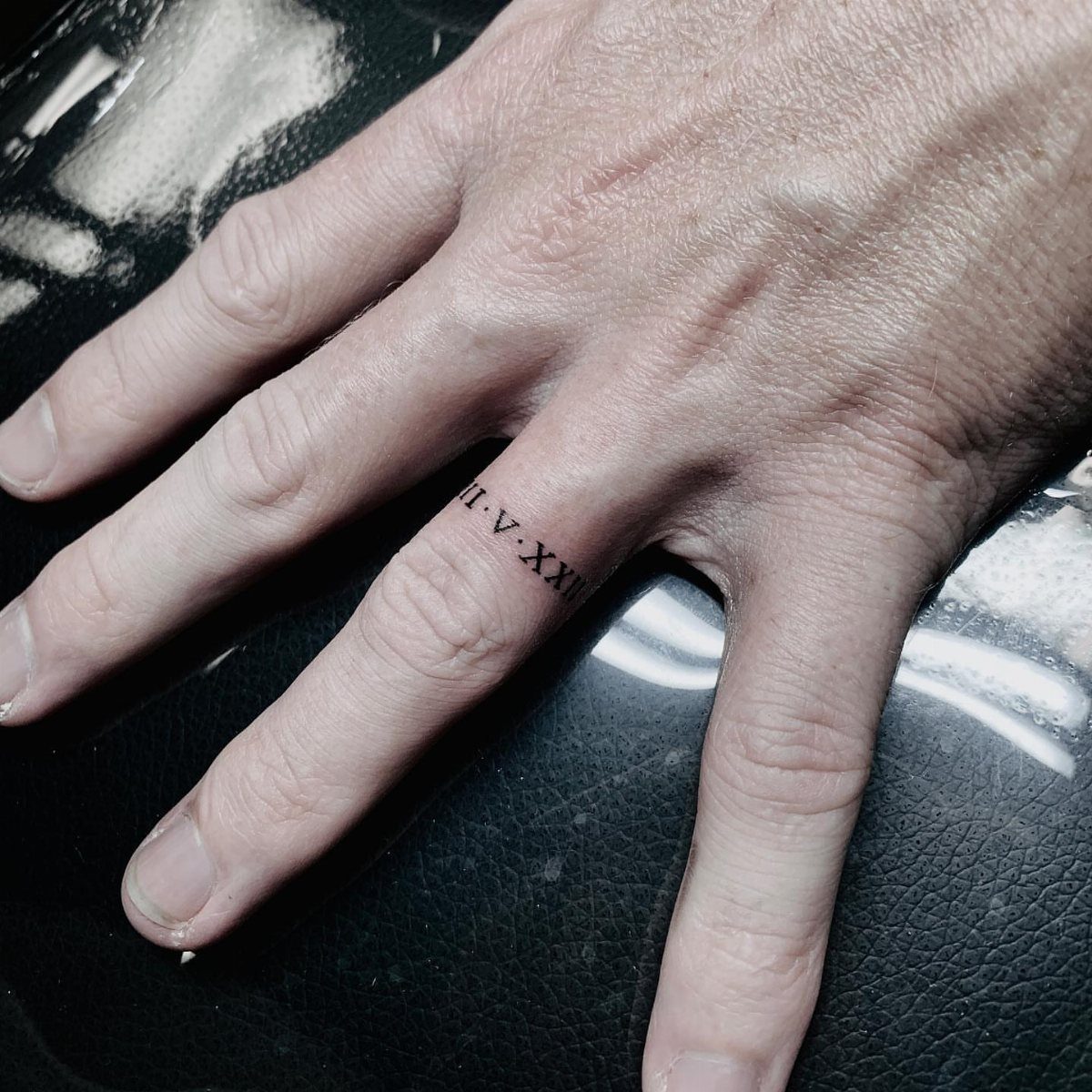 Roman numeral tattoo wrapped around a person's ring finger on a black background