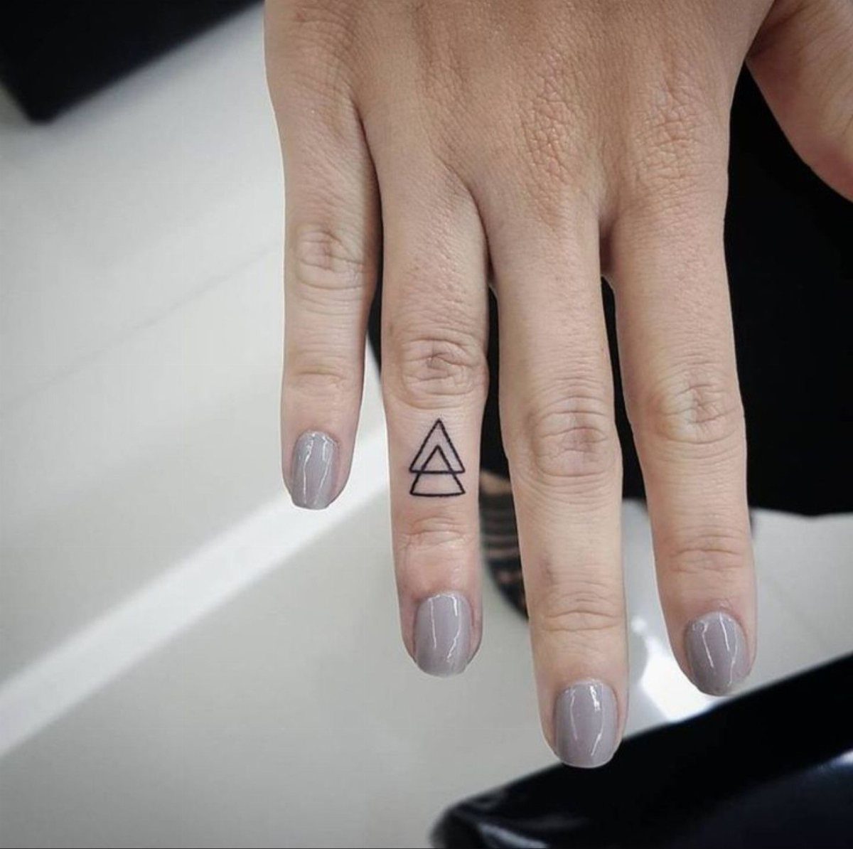 Hand with grey nails featuring two small black outline triangles tattooed on ring finger