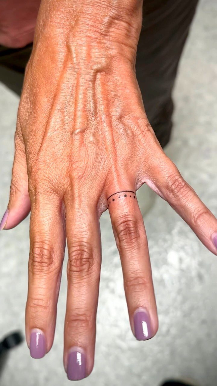 Hand with purple nails and a thin line dot tattoo on ring finger
