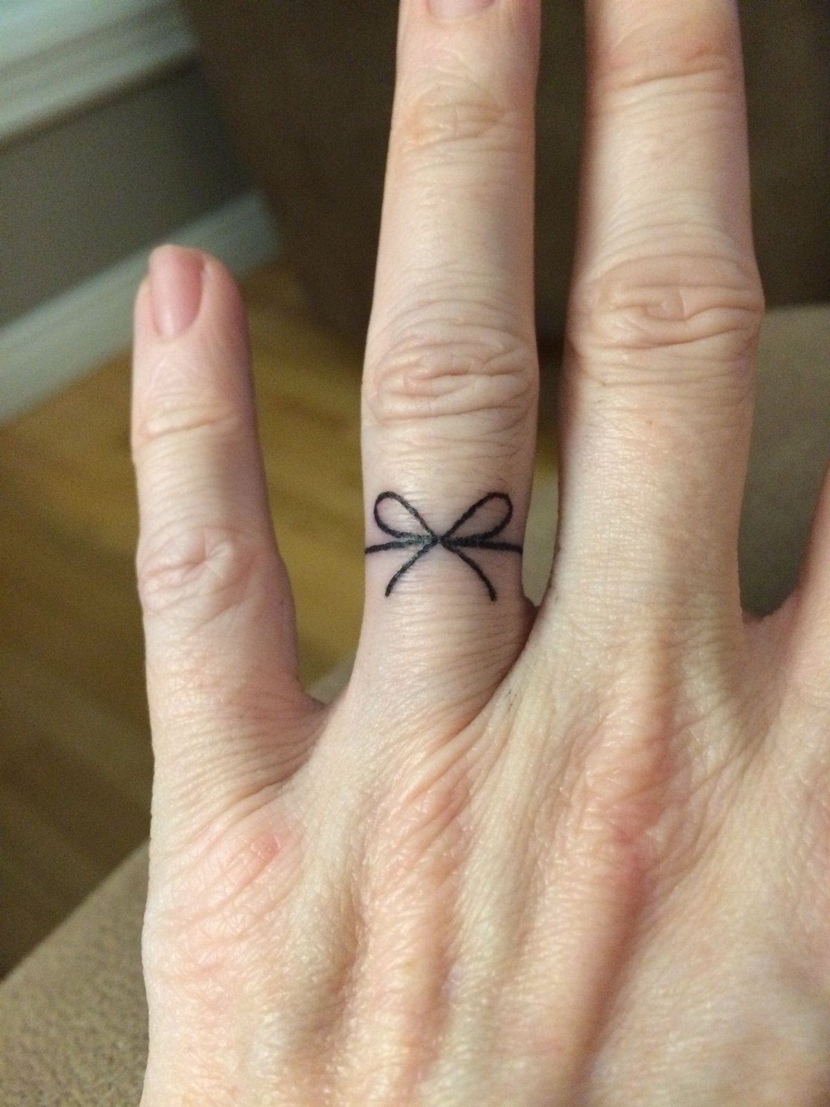 Small black ink bow tattoo wrapped around a finger like a ring.