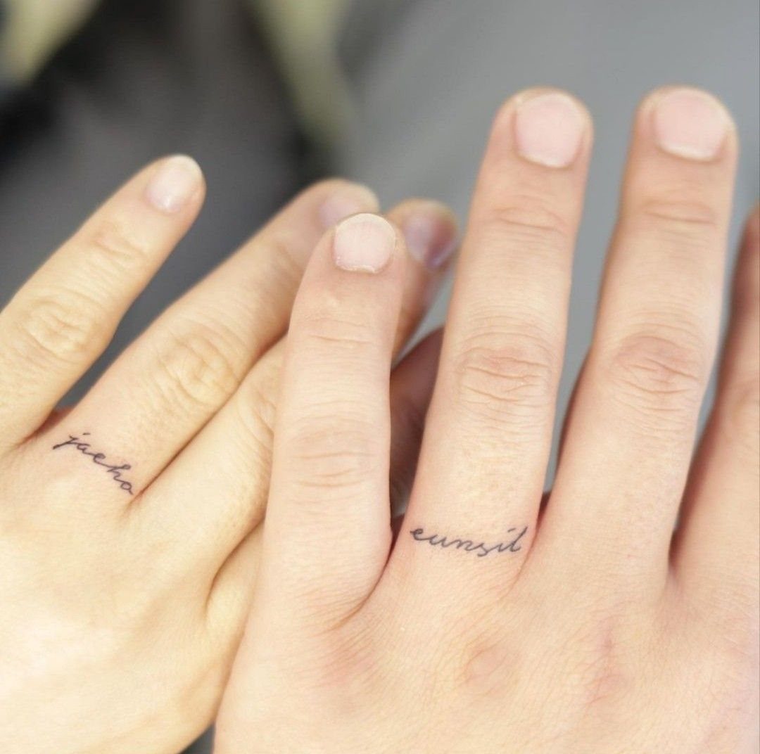 Cursive word tattoos wrapped around the ring fingers of two people's hands.