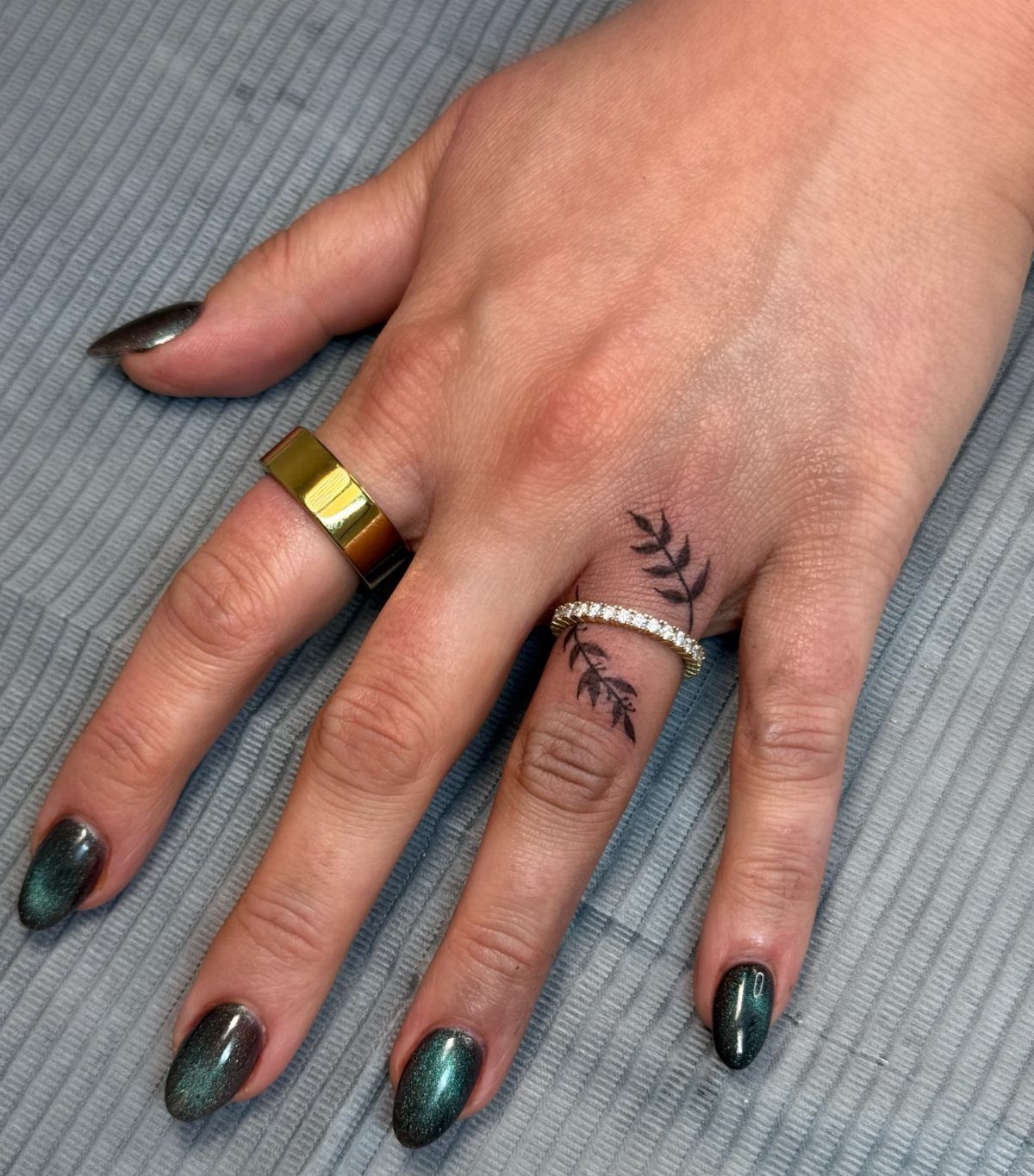 Hand with a fine-line leafy branch ring tattoo and a diamond band ring.