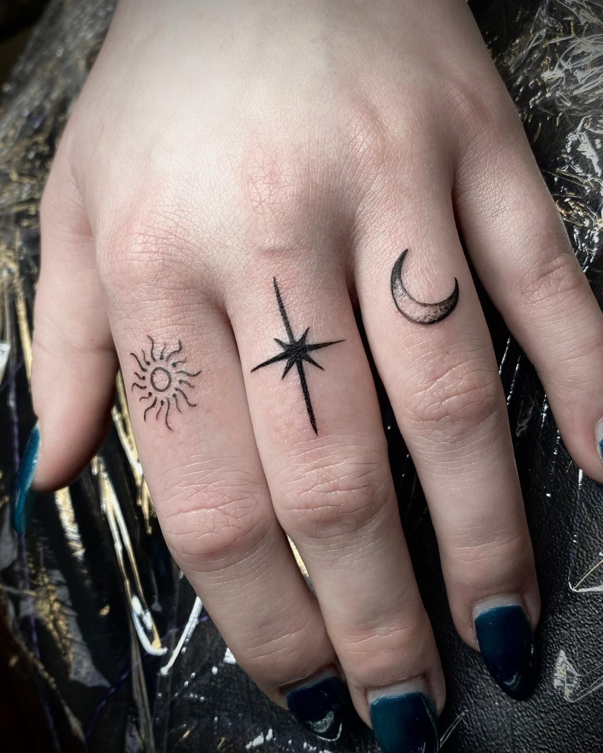 Hand with small black tattoos of a sun, star, and crescent moon on fingers