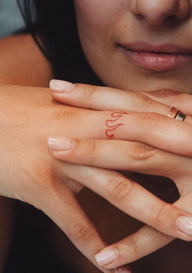 Red flame tattoo on a finger with a gold ring and manicured nails.