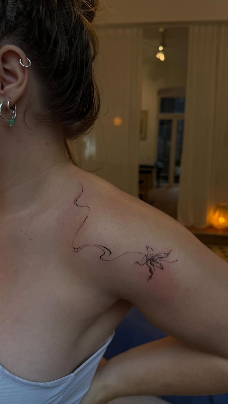 Woman's shoulder with a fine-line tattoo of a flower and a swirling stem.