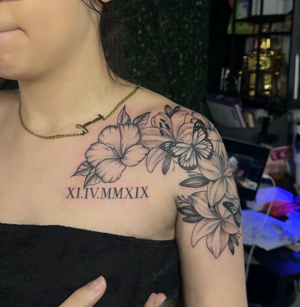Woman's shoulder with a black and grey tattoo of lilies, butterflies, and Roman numerals.