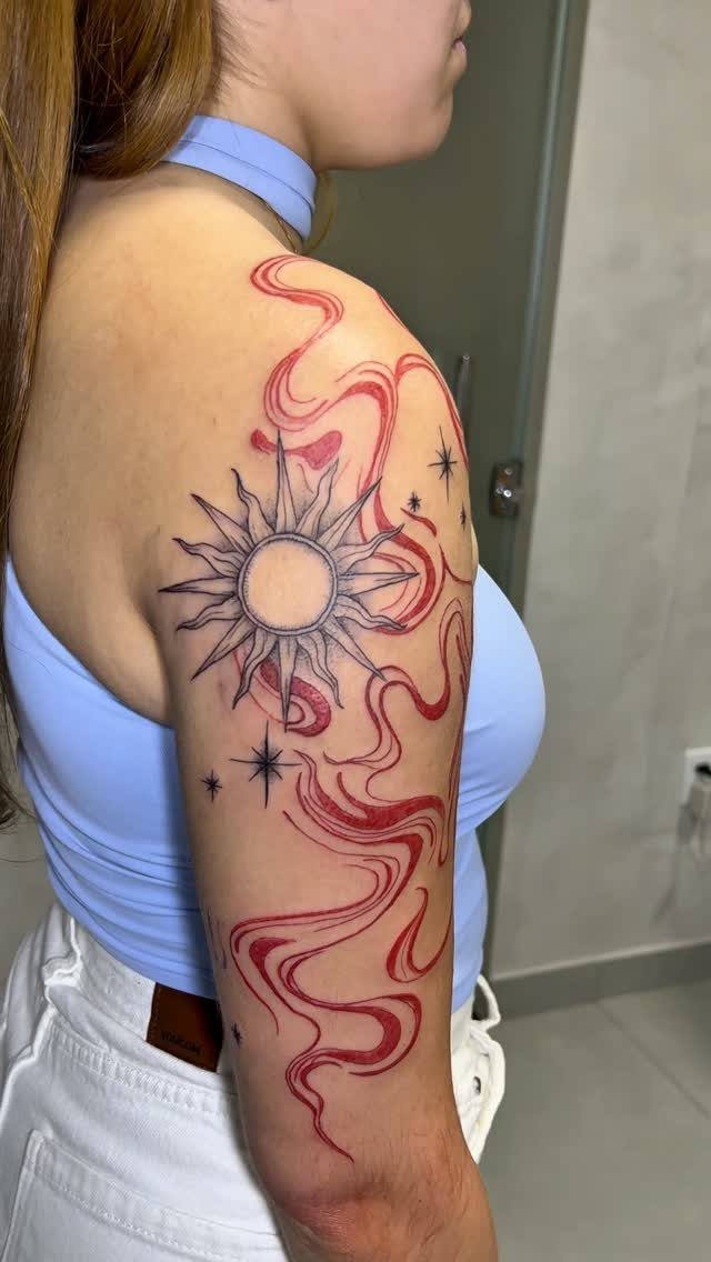 Woman's shoulder tattoo featuring a black sun with radiating spikes and flowing red lines.