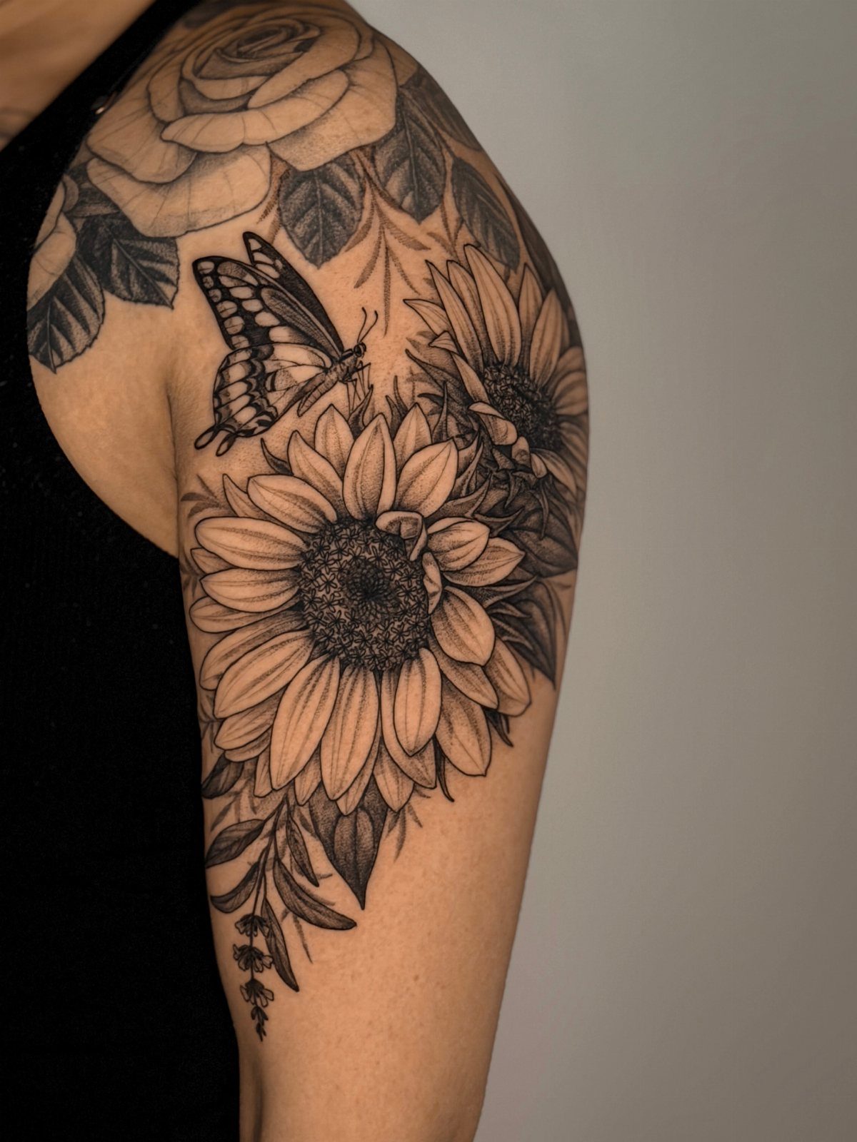 Black and grey floral tattoo on a shoulder featuring sunflowers and a butterfly.