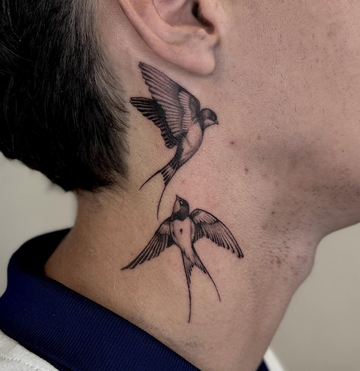 Two black swallow bird tattoos on the side of a person's neck.