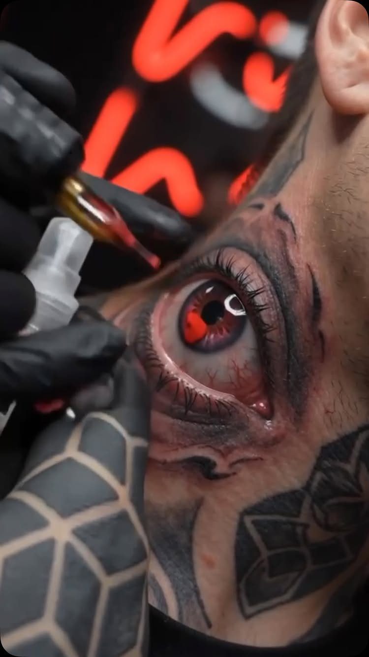 Realistic red eye tattoo on a person's neck being treated by a tattoo artist.