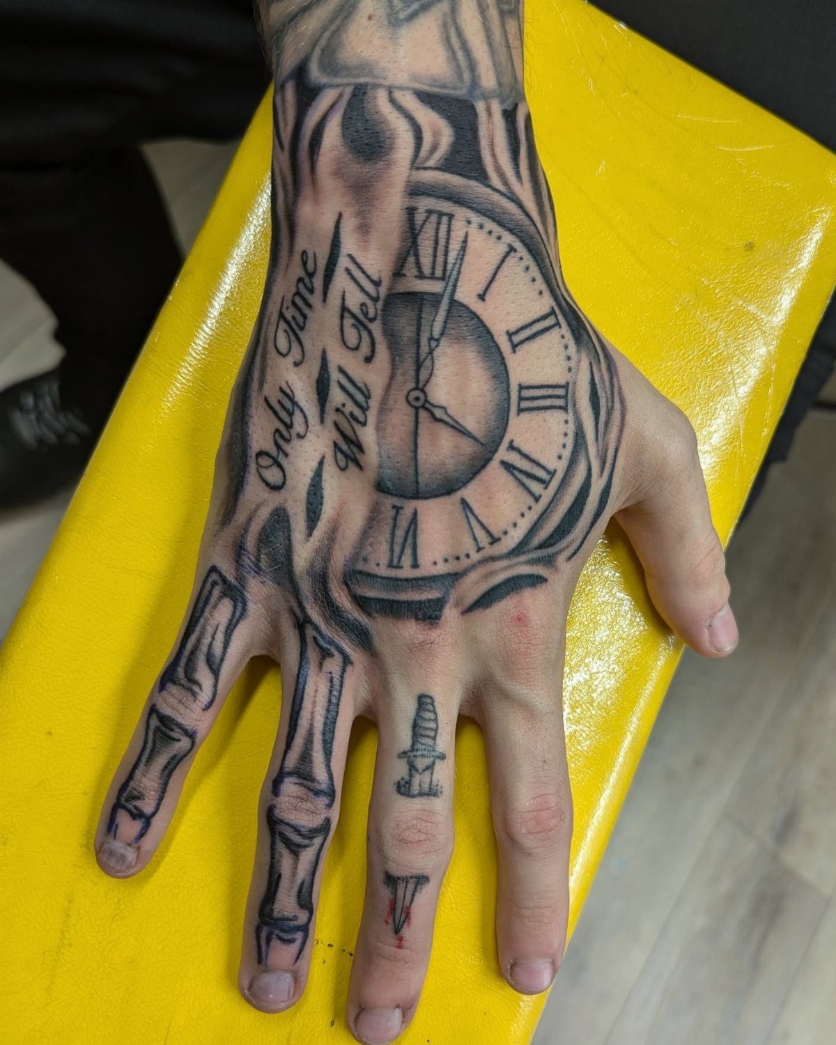 Hand tattoo featuring a Roman numeral clock and skeletal finger bones on skin.