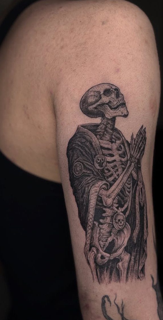 Skeleton tattoo on an arm wearing a cloak with hands in prayer position.
