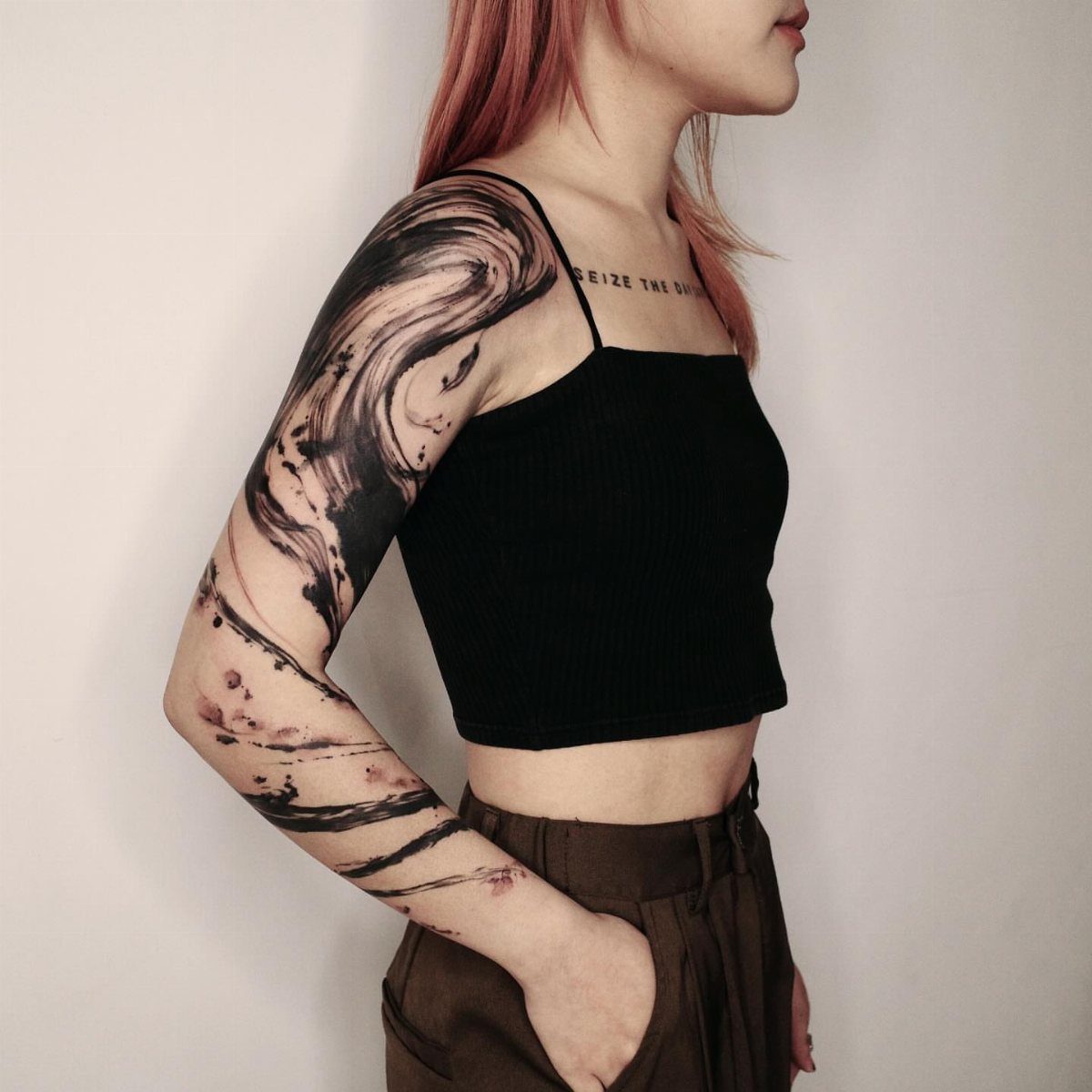 Woman with pink hair showing a black abstract brushstroke sleeve tattoo on her arm.