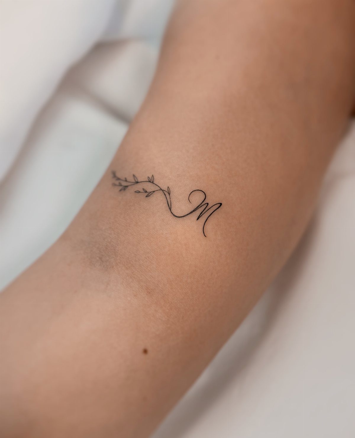 Cursive letter M tattoo with a small leafy branch on a person's wrist