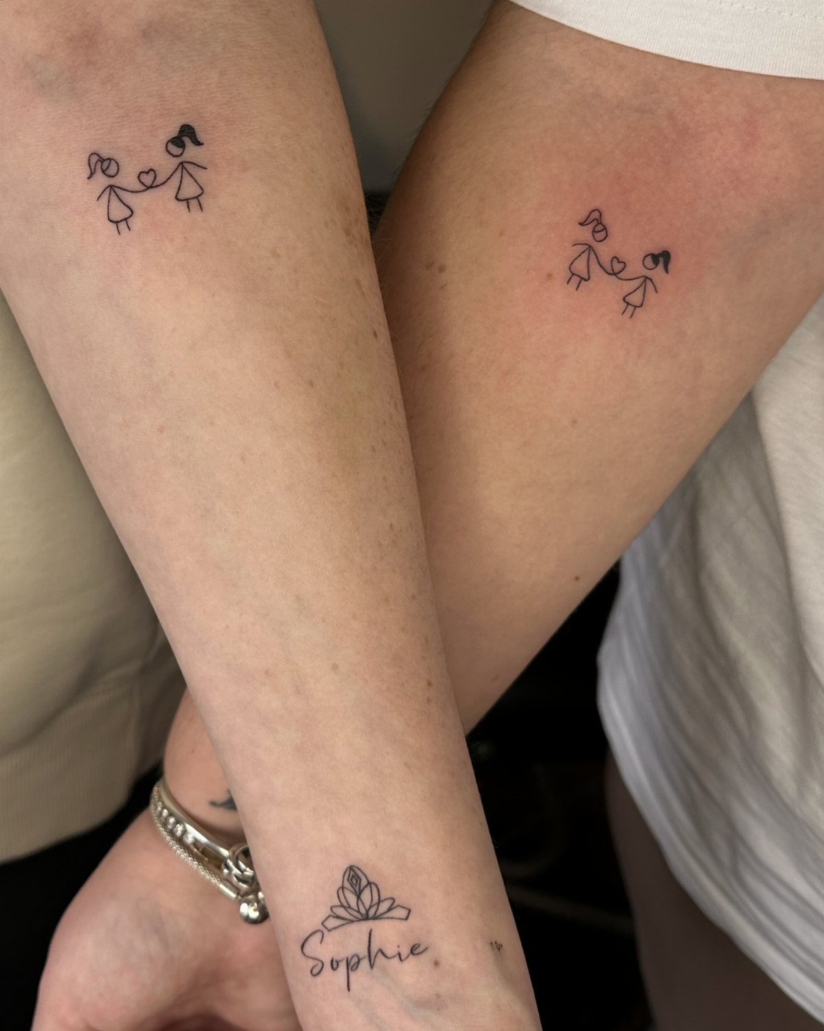 Two matching tattoos of stick-figure girls holding hands on two different arms.