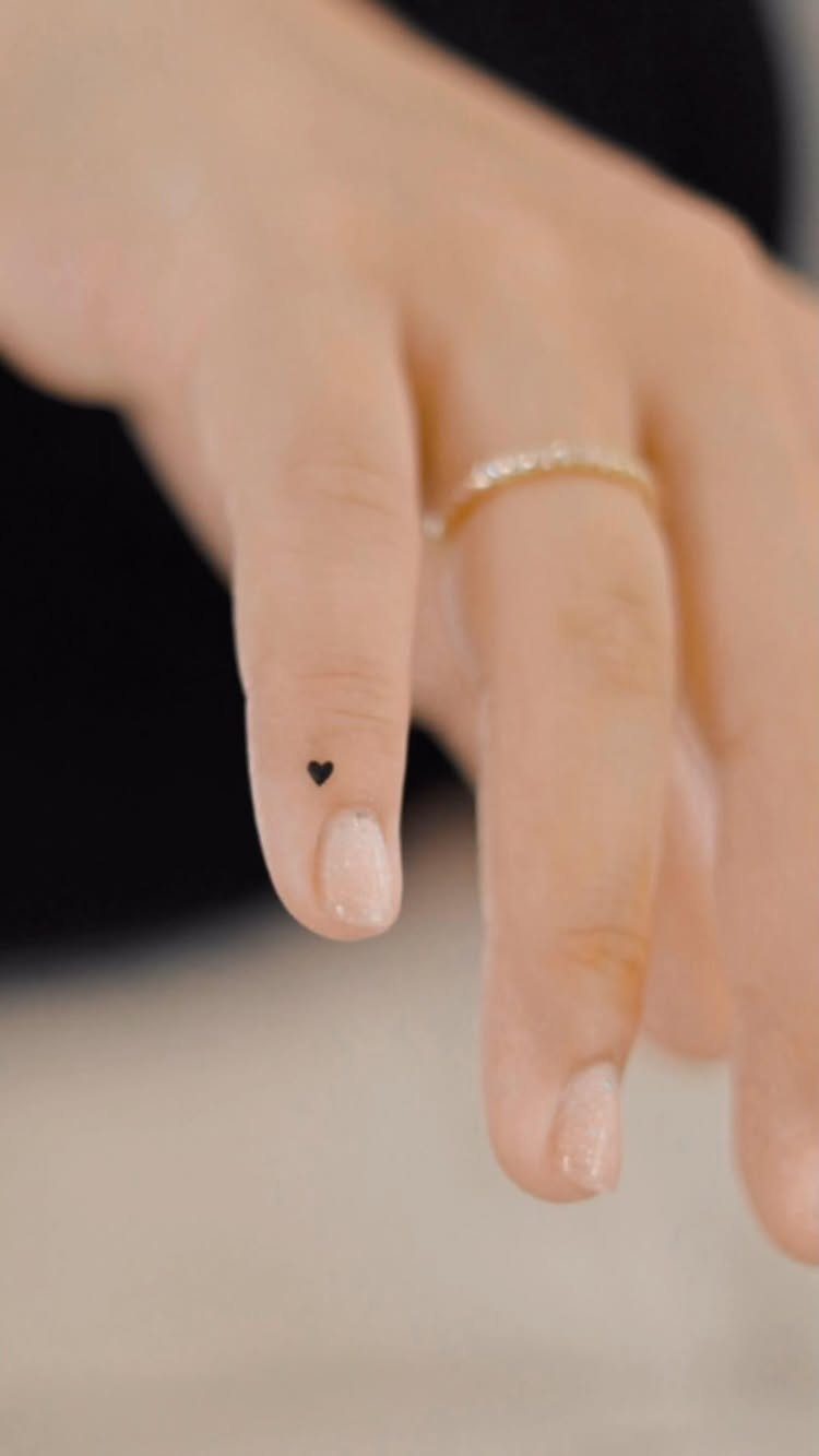 Tiny black heart tattoo on the side of a finger with a gold ring