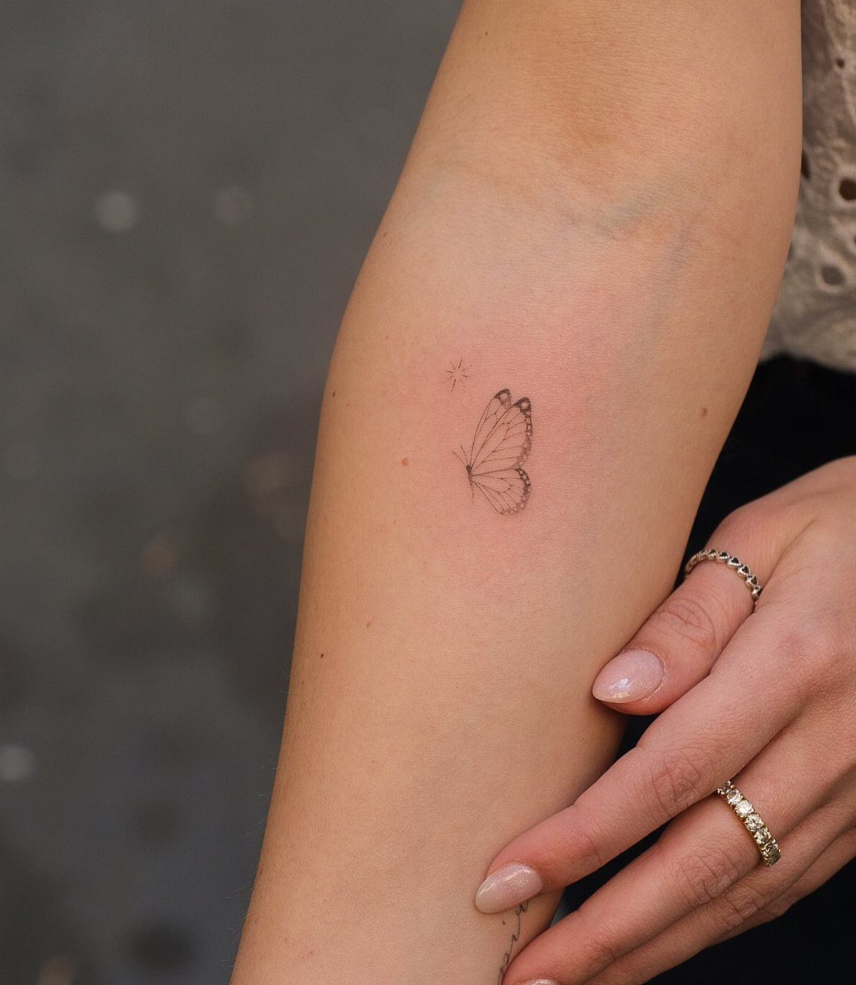 Small delicate butterfly tattoo on a person's forearm with silver rings and manicured nails.