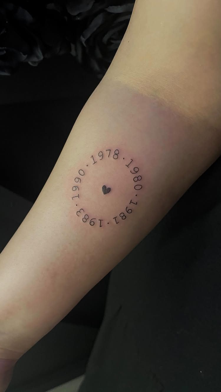 Small heart tattoo surrounded by a circle of birth years on a forearm