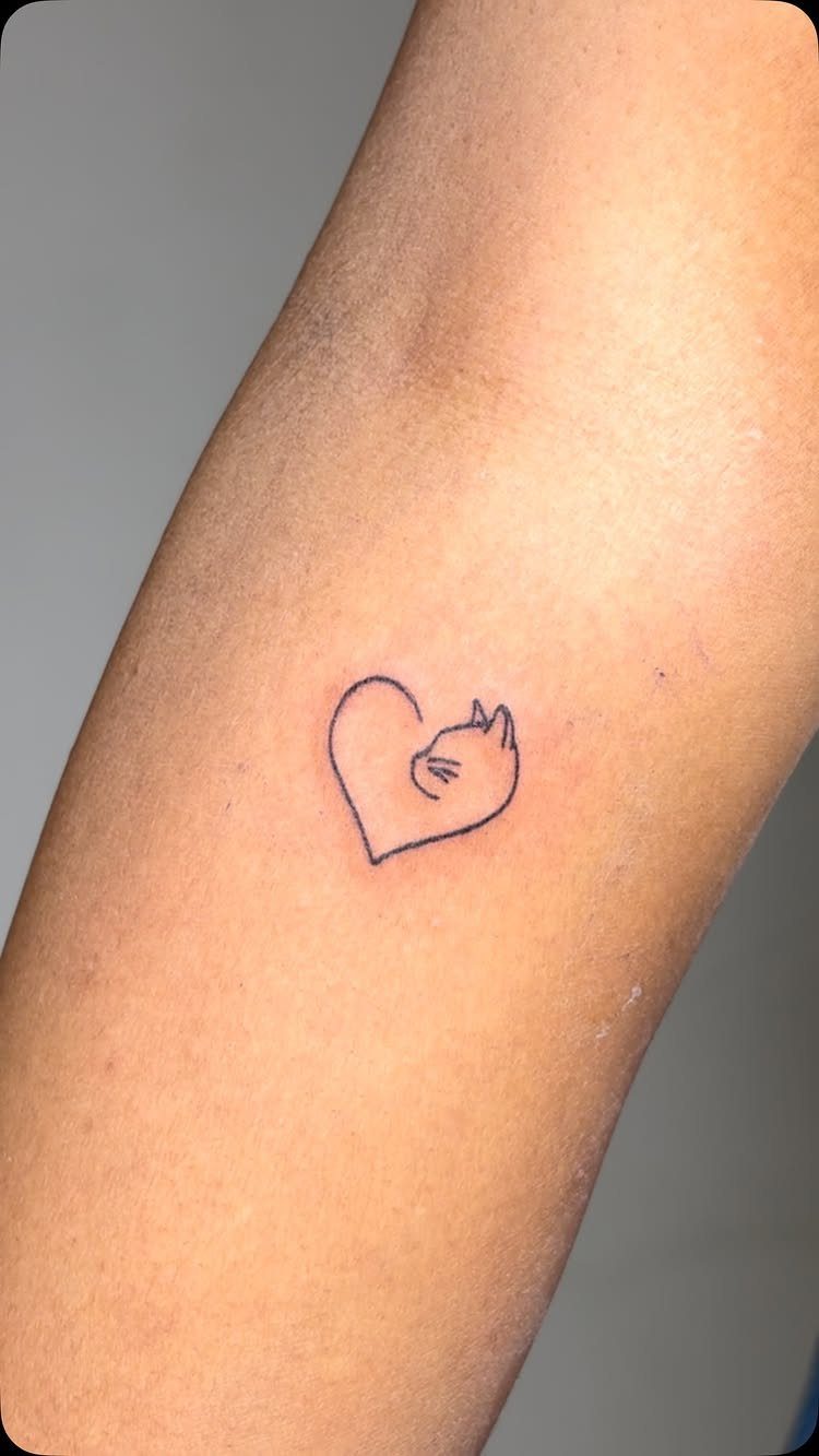 Small black ink tattoo of a cat's head forming a heart on an arm