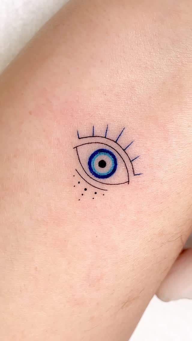 Small eye tattoo with a blue iris and black outlines on fair skin.