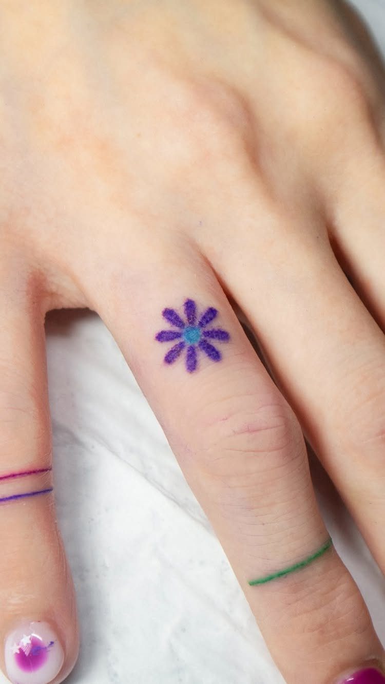 Small purple flower tattoo on a finger with thin colored band tattoos.