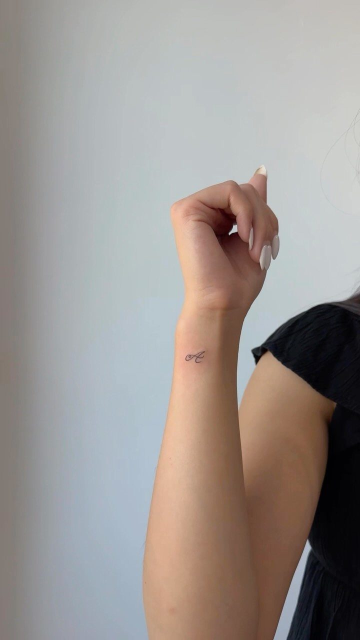 Small cursive letter tattoo on a person's inner wrist with white nails.