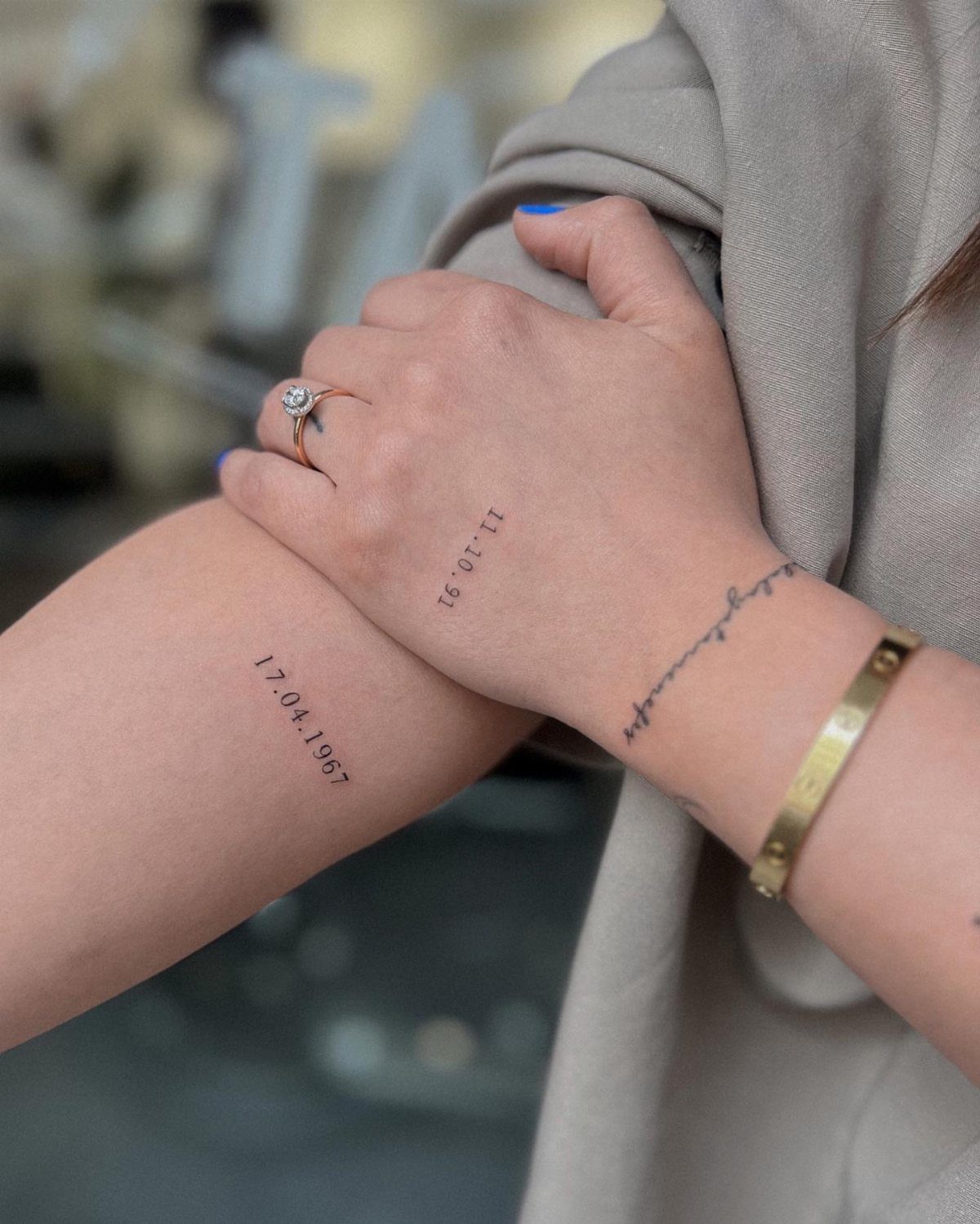 Two wrists featuring small black ink tattoos of meaningful dates and a gold ring.