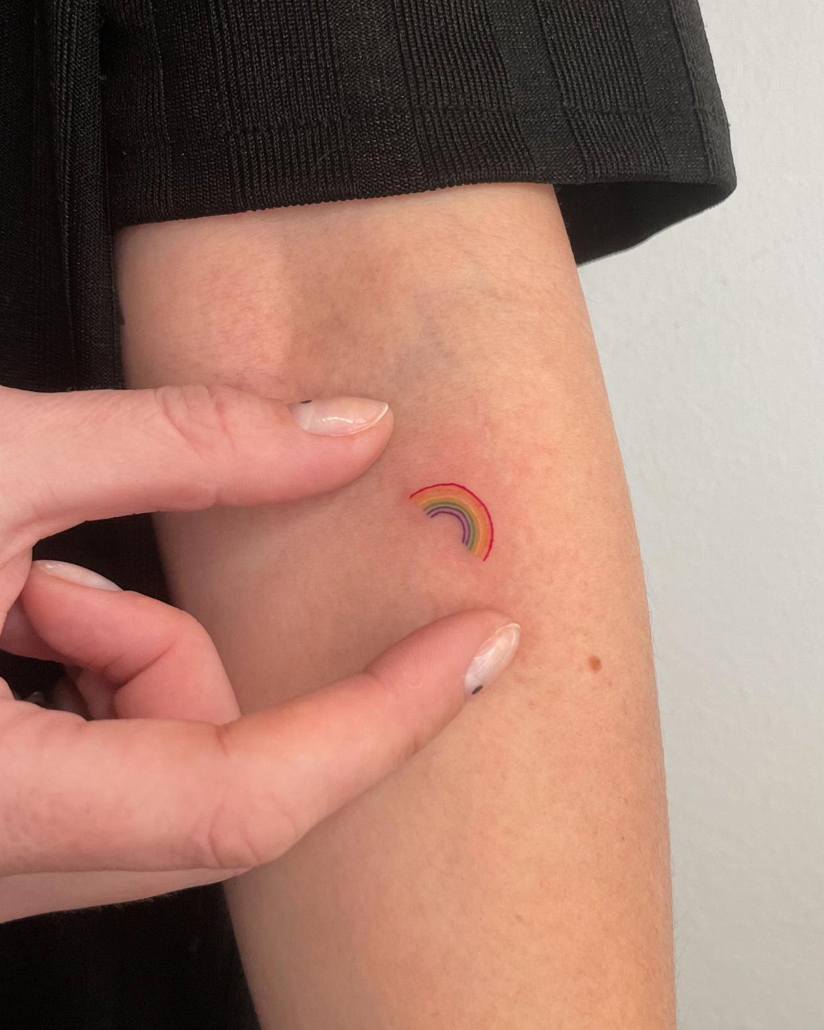 Small colorful rainbow tattoo on a person's upper arm being pointed to by fingers