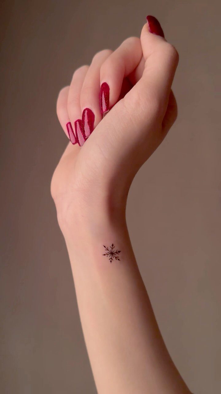 Small black snowflake tattoo on a person's inner wrist with red nails.