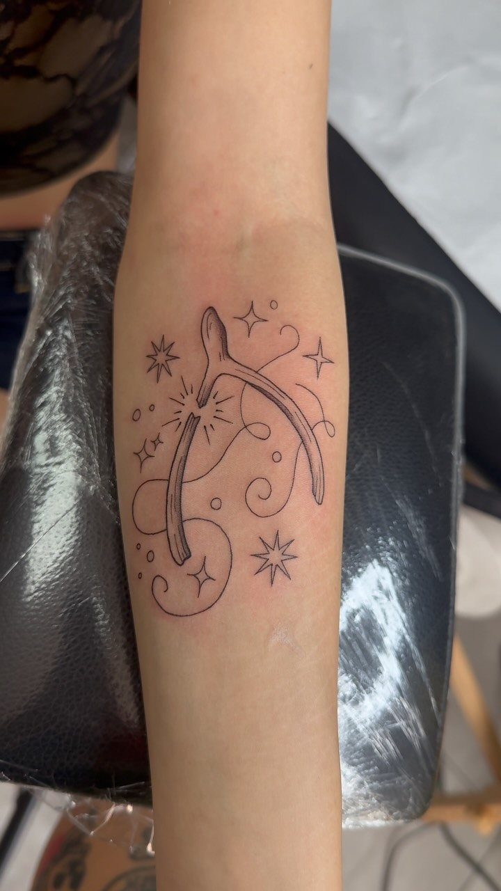 Wishbone tattoo on a forearm with small stars and decorative swirls in black ink.