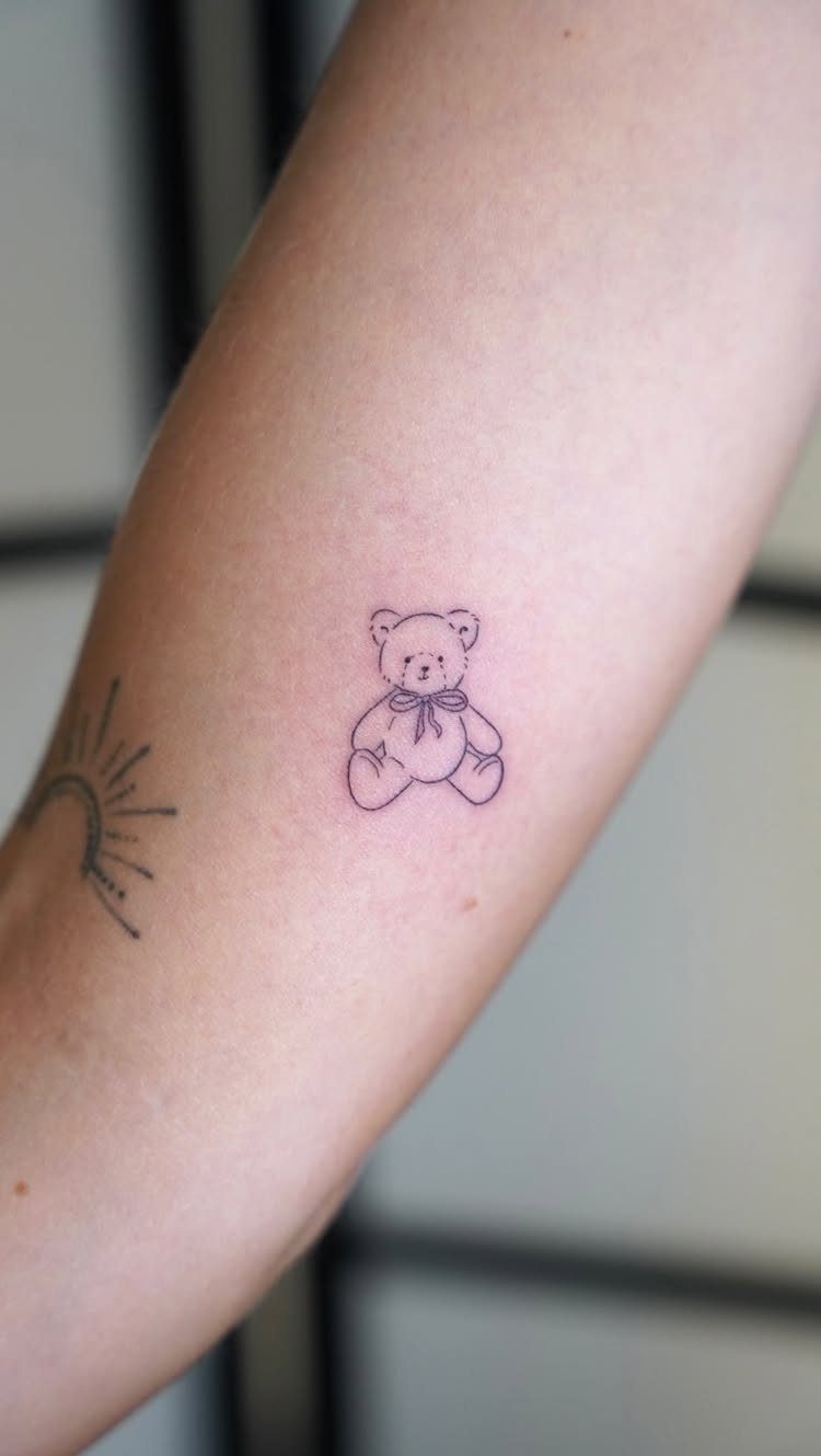 Small teddy bear tattoo with a bow on a person's forearm.