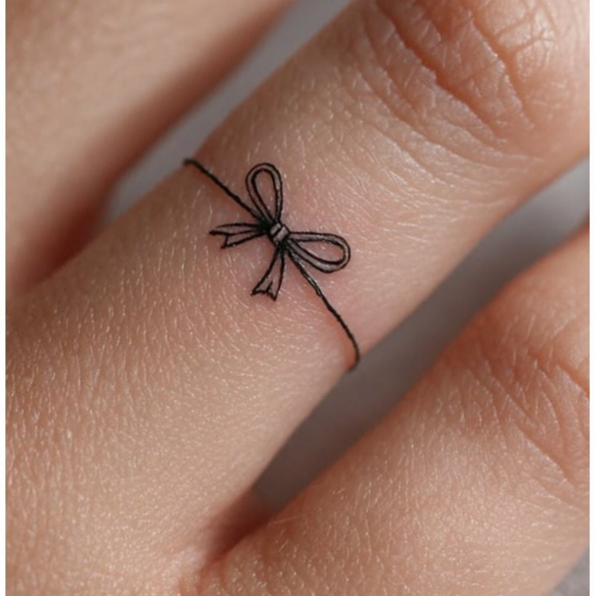Small black ink bow tattoo encircling a finger like a ring.