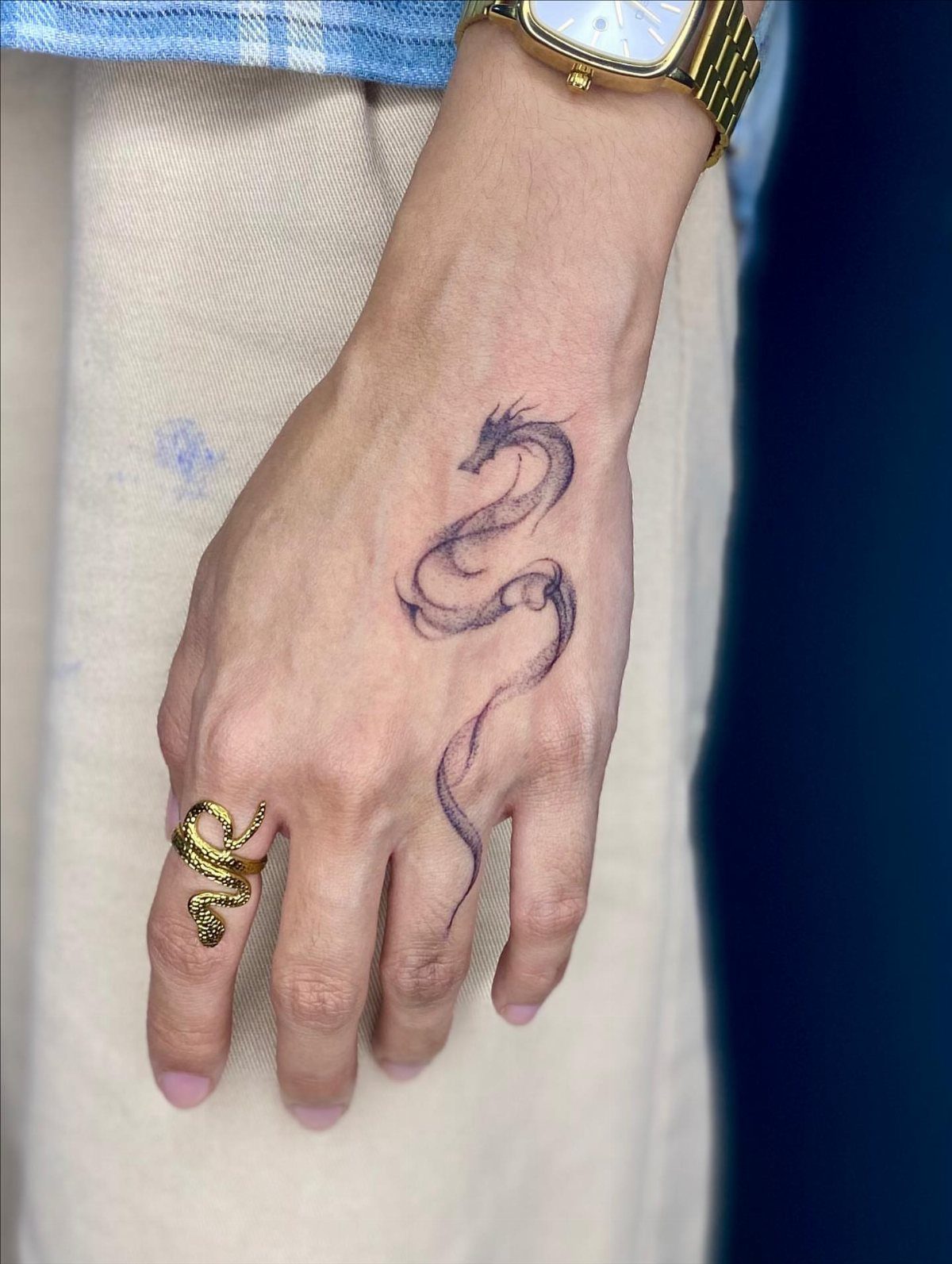 Slender black dragon tattoo on the back of a hand with gold jewelry.
