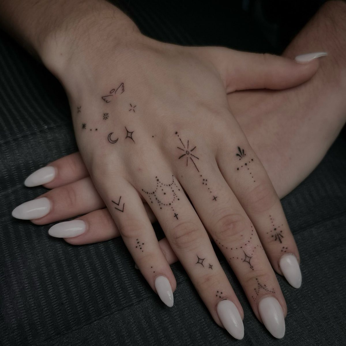 Hands with white nails featuring small sun, moon, and star tattoos.