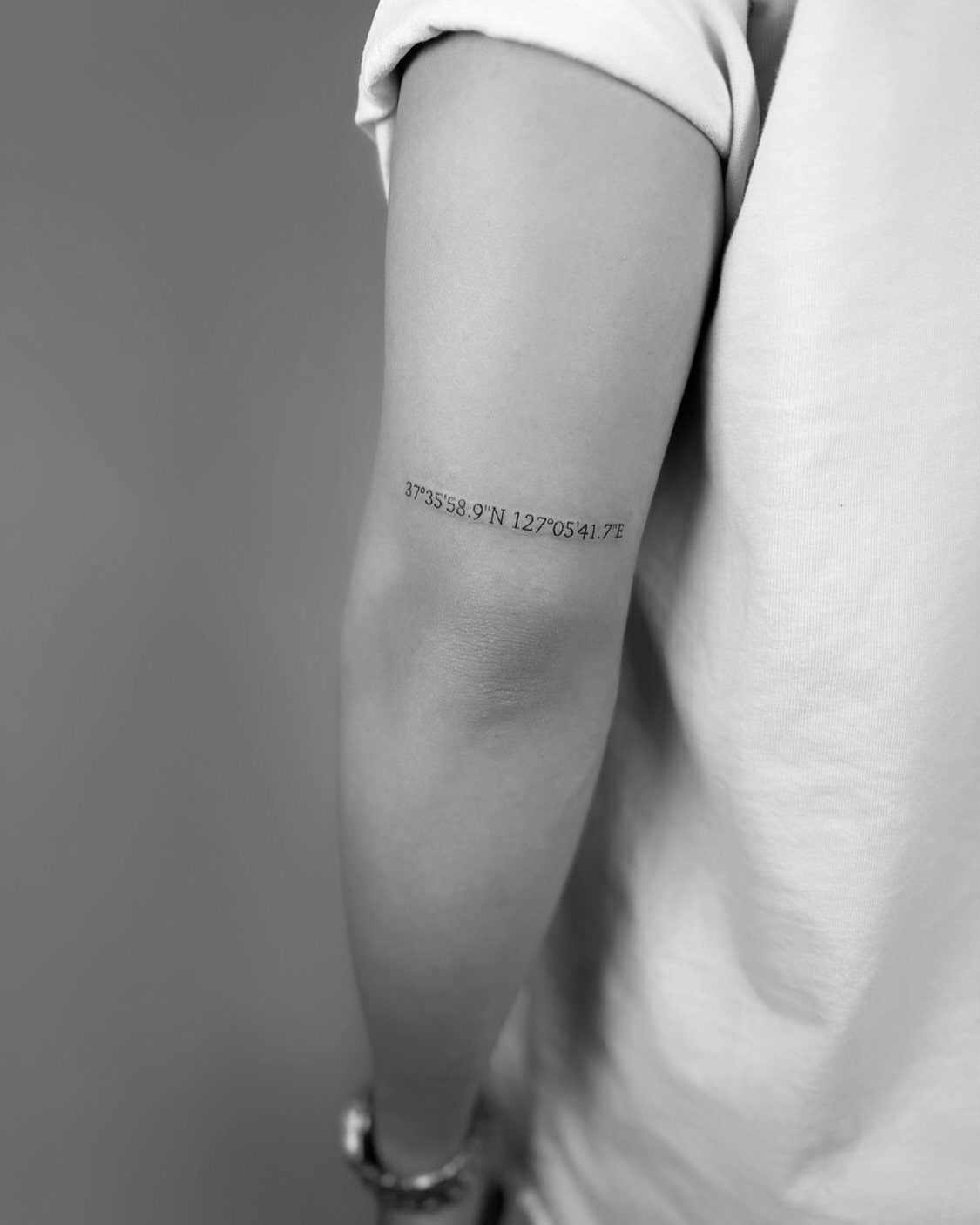 Small coordinate tattoo on a person's upper arm in black and white.