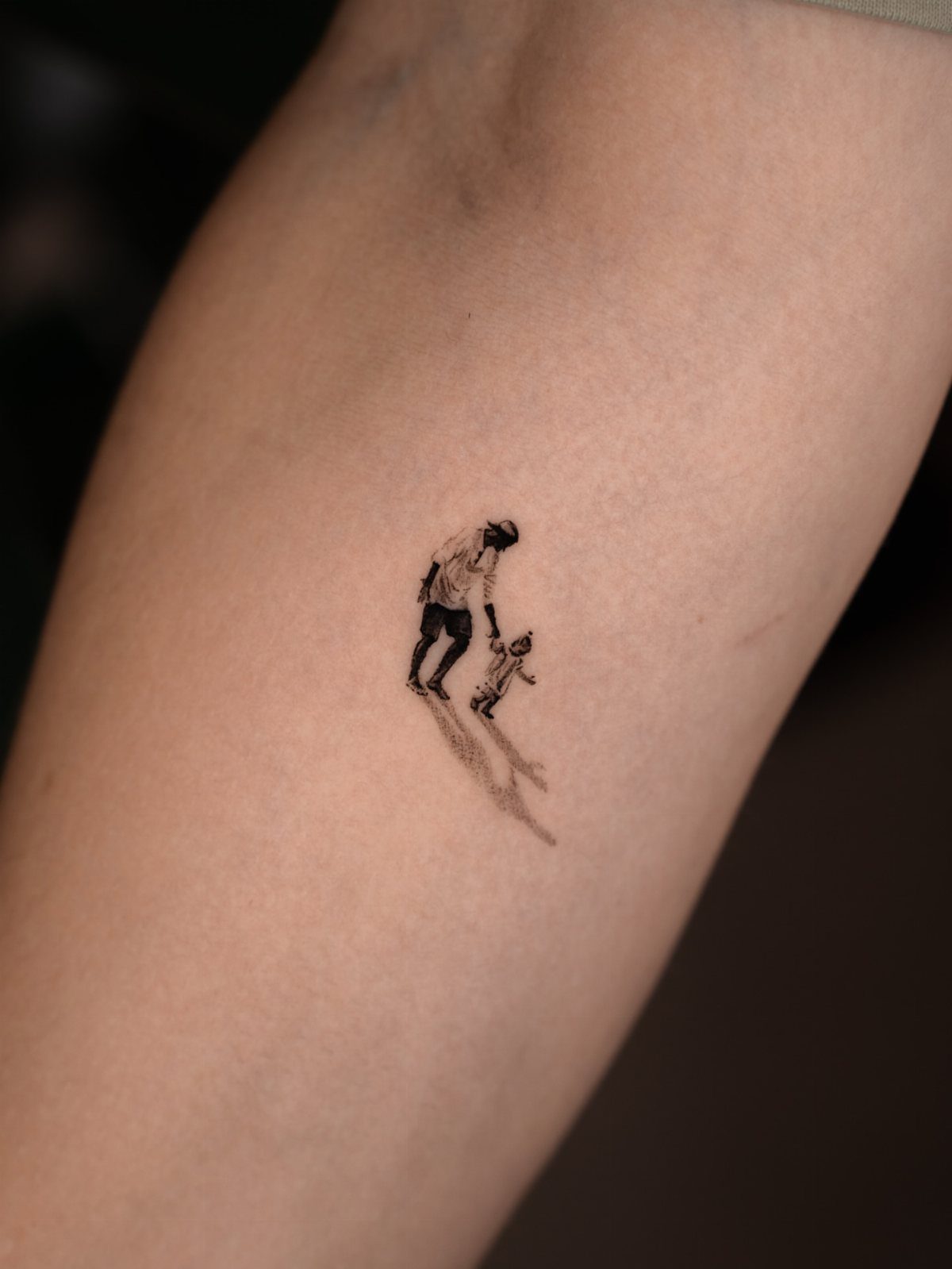Small black ink tattoo of a man and child holding hands on an arm