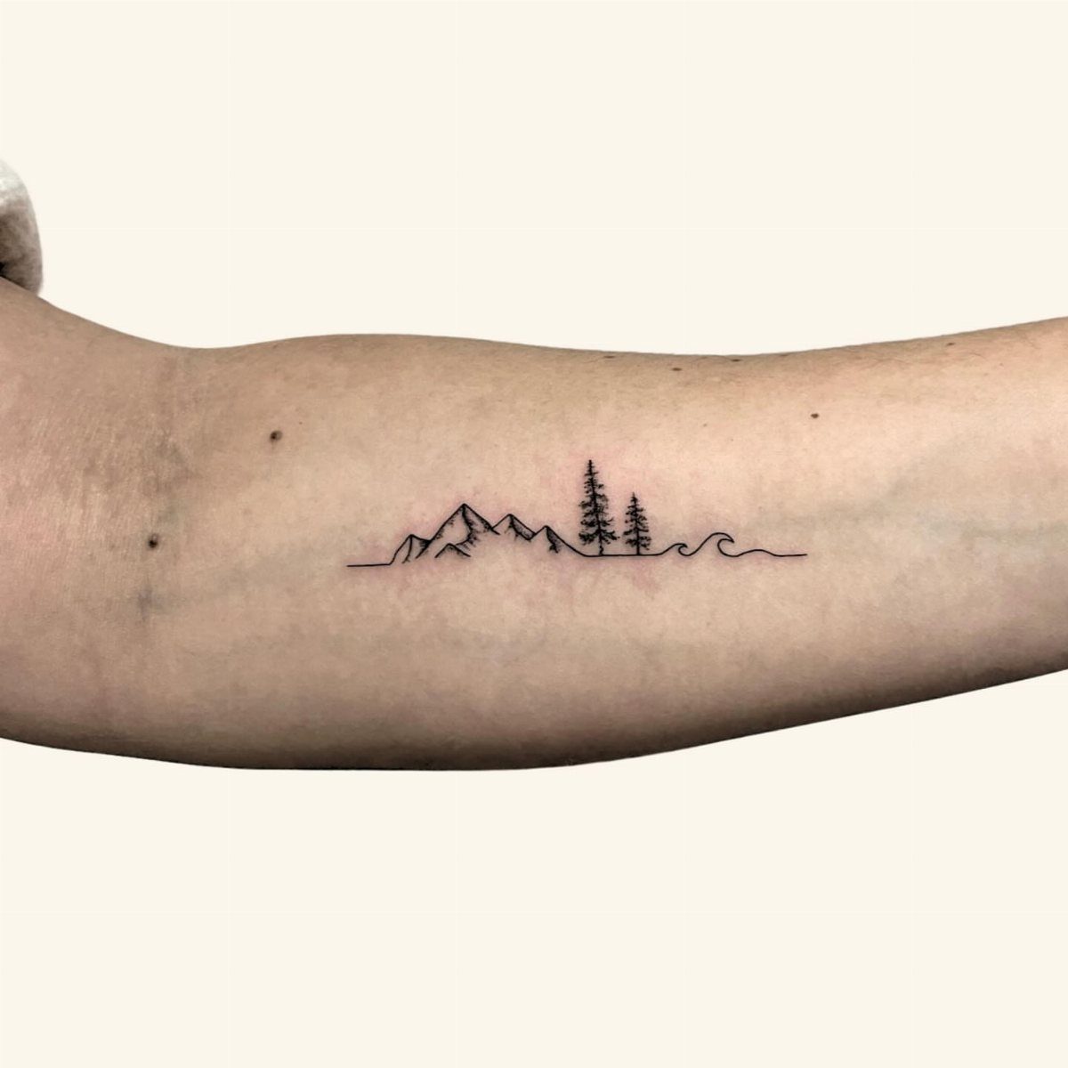 Mountain silhouettes and pine trees tattooed in fine black lines on a forearm.